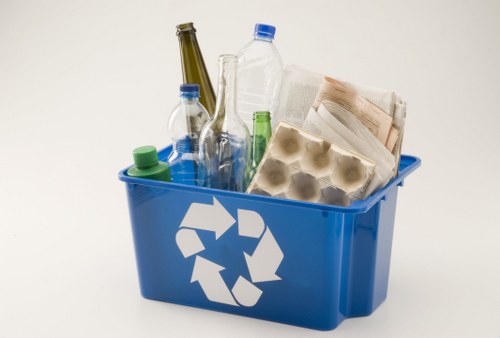 A blue recycling bin with a white recycling symbol on the front, filled with various waste items including plastic bottles, glass bottles, newspapers, and paper egg cartons, situated on a plain light background. The plastic bottles are of different sizes and colors, such as green and clear, with some still capped. The glass bottles are taller, with one appearing to be a wine bottle with a dark green hue. The newspapers are folded and placed on top of the other waste, showing visible text and images. The paper egg cartons are made of recycled cardboard and have a light brown color. The bin appears to be positioned in an indoor or studio setting, emphasizing the importance of proper rubbish separation and waste management, which is relevant to professional rubbish removal services like those offered by Office Clearance Crouch End in the local area.