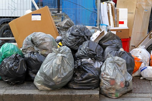 A collection of tightly tied black and gray plastic rubbish bags filled with waste material, placed on a paved surface likely on a pavement or driveway in an urban area. Several bags are piled together, some leaning against each other, with a few partially transparent bags revealing mixed general refuse inside. In the background, there are cardboard boxes, a red plastic container, and other miscellaneous items, indicating a rubbish clearance or disposal scene typical of a commercial or residential street in or near Crouch End. The bags are positioned at the edge of the pavement, and the environment appears to be a typical waste collection or rubbish removal setup, consistent with services offered by Office Clearance Crouch End in the local area. The lighting suggests natural daylight, highlighting the textures of the plastic and cardboard materials.
