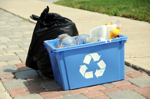 A blue plastic recycling bin prominently positioned on a brick-paved sidewalk surface, featuring a white recycling symbol on its side. The bin is filled with clear plastic bottles, including some with labels visible, as well as a few discarded containers. Adjacent to the bin, on the left, there is a tied black garbage bag placed on the ground, leaning slightly against the bin. The surrounding environment includes a paved path leading into a grassy area with a blurred background of green lawn and part of a suburban street scene, indicating a typical outdoor rubbish collection setting in Crouch End. The scene appears well-lit with natural daylight, highlighting the different textures and colors of the waste materials, the bin, and the pavement, consistent with a professional rubbish removal service context performed by Office Clearance Crouch End.