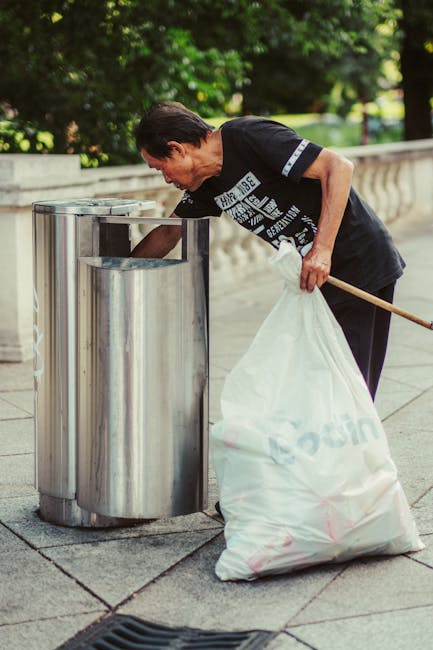A black wheeled rubbish bin positioned on a sidewalk in an urban area during nighttime, with the label 'ST. JOHN'S' on the front. The bin is partially open, revealing various waste items, including wh