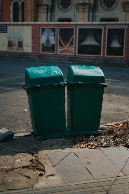 A dark alleyway cluttered with large bags and overflowing rubbish, including cardboard boxes, black garbage bags, and various discarded items stacked against a makeshift fence at the rear. The ground