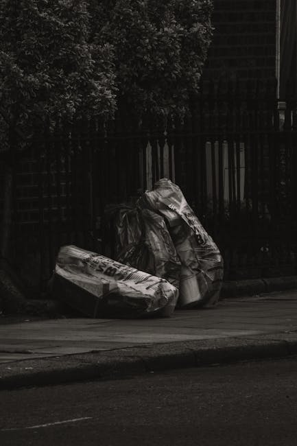 An outdoor scene showing a large pile of black, white, and red rubbish bags, some partially torn open, filled with various discarded packaging, paper, and household waste, situated next to recycling c