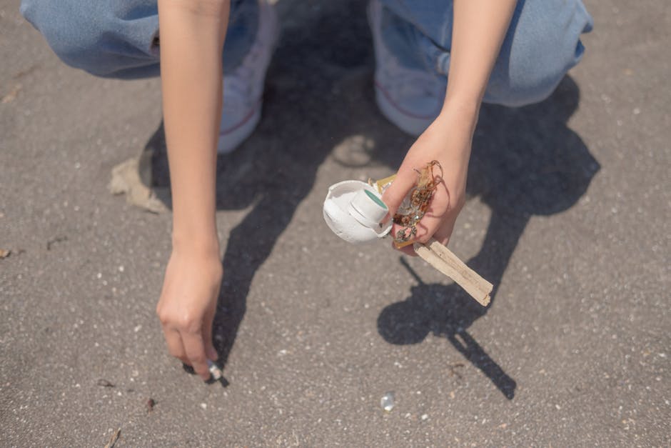 A person crouching on a paved outdoor surface, wearing light blue jeans and white sneakers, is holding a small, broken white ceramic object attached to a wooden stick. The ceramic piece has a smooth,