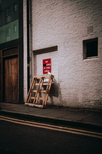 A pile of black rubbish bags and discarded packaging materials, including a cardboard box and foam padding, stacked in front of a red metal fire door on a beige exterior wall. The door is covered with