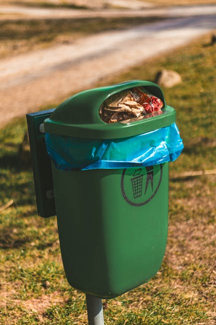 A residential street scene showing a row of dark green wheelie bins with white painted numbers lined up along the pavement outside brick houses. A woman in a brown coat stands near one of the bins, po